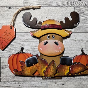May include: A wooden moose decoration with a straw hat, sunglasses, and two orange pumpkins. The moose is surrounded by fall leaves and has a tag that says "MOOSE FALL". The decoration is on a wooden background.