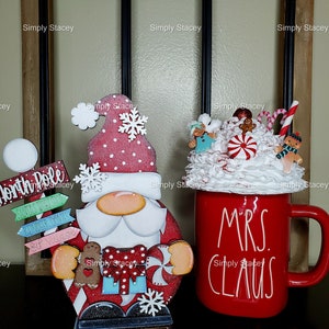 May include: A red and white wooden Santa Claus figurine with a white beard and a red hat. The figurine is holding a candy cane and a gingerbread man. The figurine is standing on a wooden base with the words "North Pole" and "Santa's Workshop" written on it. There is also a red mug with white lettering that says "Mrs. Claus".