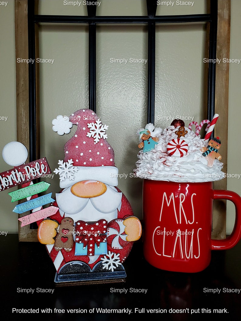 May include: A red and white wooden Santa Claus figurine with a white beard and a red hat. The figurine is holding a candy cane and a gingerbread man. The figurine is standing on a wooden base with the words "North Pole" and "Santa's Workshop" written on it. There is also a red mug with white lettering that says "Mrs. Claus".