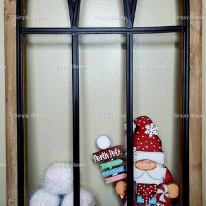 May include: A red and white gnome with a white beard and a red hat with a snowflake on it. The gnome is holding a sign that says "North Pole". The gnome is in front of a black metal window frame with white fluffy balls in the bottom left corner.
