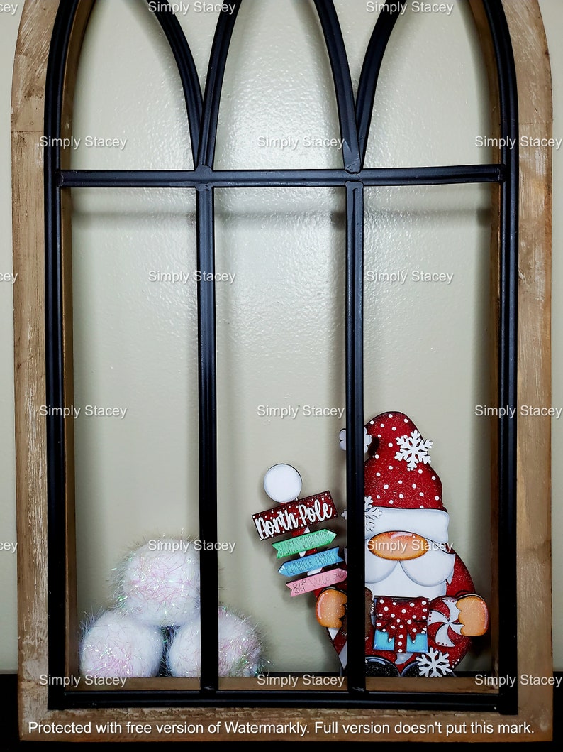 May include: A red and white gnome with a white beard and a red hat with a snowflake on it. The gnome is holding a sign that says "North Pole". The gnome is in front of a black metal window frame with white fluffy balls in the bottom left corner.