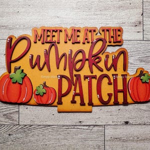 May include: Wooden sign with the text "MEET ME AT THE PUMPKIN PATCH" in red and orange lettering. The sign features three orange pumpkins with green leaves. The background is a light wood grain.
