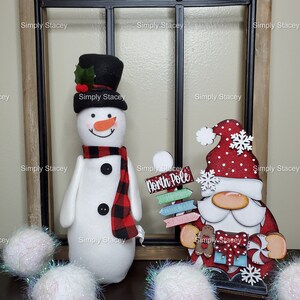 May include: A white snowman with a black top hat and red and black plaid scarf stands next to a red and white Santa Claus figure holding a sign that says "North Pole". Both figures are in front of a black metal window frame. Three white pom-poms are in the foreground.