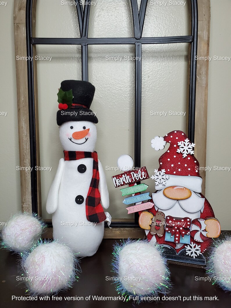 May include: A white snowman with a black top hat and red and black plaid scarf stands next to a red and white Santa Claus figure holding a sign that says "North Pole". Both figures are in front of a black metal window frame. Three white pom-poms are in the foreground.