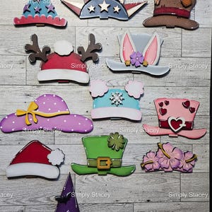 May include: A collection of decorative wooden hat cutouts in various designs, including a patriotic crown, a reindeer hat, a bunny ear hat, a polka dot hat, a snowflake hat, a heart hat, a Santa hat, a leprechaun hat, a floral hat, and a witch hat.