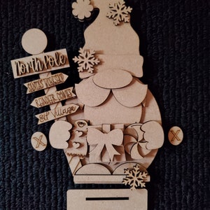 May include: Wooden craft kit featuring a gnome holding a gift and candy cane. The kit includes signs that say "North Pole", "Santa's Workshop", "Reindeer Games", and "Elf Village".