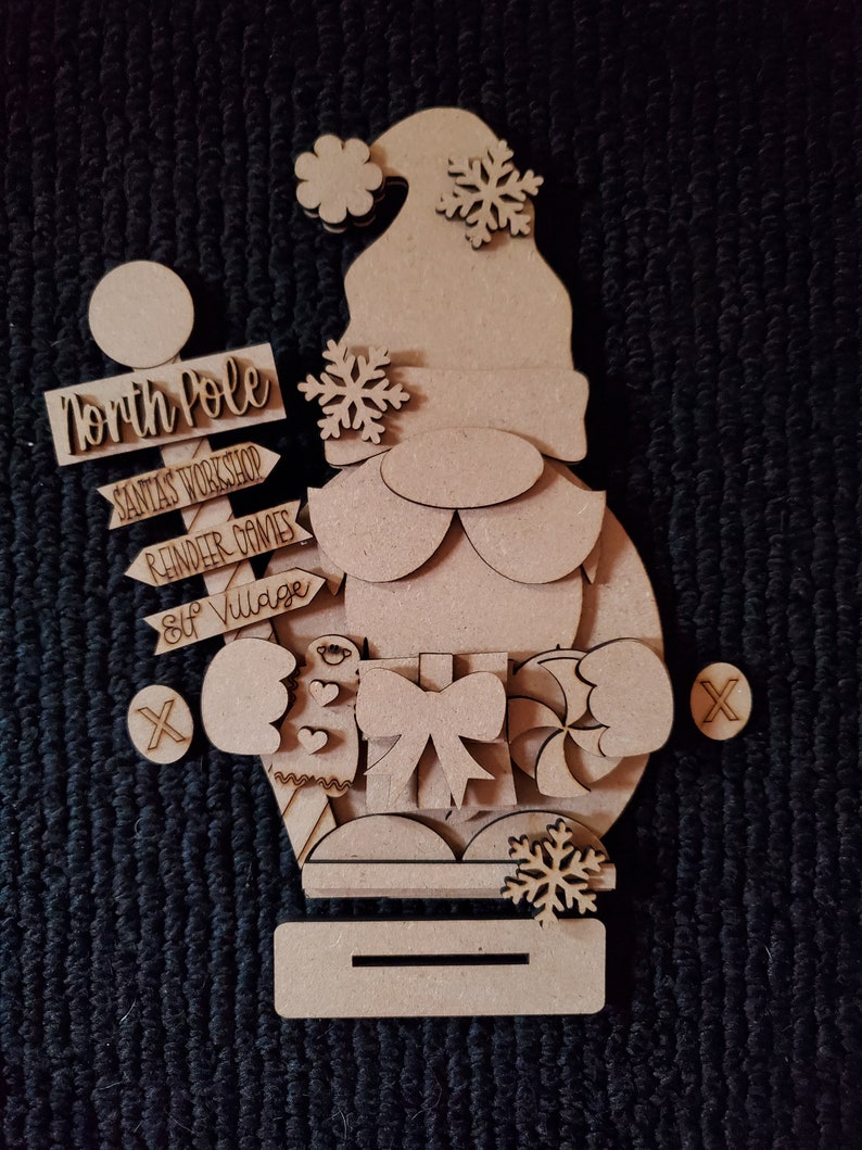 May include: Wooden craft kit featuring a gnome holding a gift and candy cane. The kit includes signs that say "North Pole", "Santa's Workshop", "Reindeer Games", and "Elf Village".