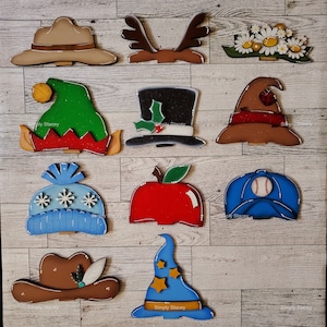May include: Twelve different wooden cutout shapes painted in various colors and designs. The shapes include a cowboy hat, a reindeer, a flower crown, an elf hat, a top hat, a witch hat, a winter hat, an apple, a baseball cap, a cowboy hat with a feather, a wizard hat, and a cowboy hat with a feather.