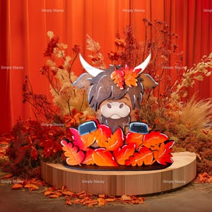 May include: A wooden cutout of a Highland cow with a brown and white face, wearing a crown of fall leaves. The cow is standing on a wooden base surrounded by fall leaves.