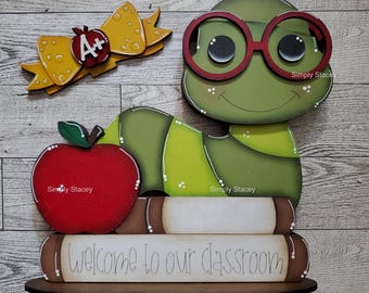 May include: Wooden classroom decoration featuring a green turtle wearing red glasses, a red apple, and a yellow bow with an apple and the letter "A+". The words "Welcome to our classroom" are written on a stack of books.