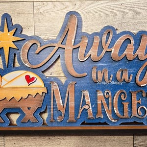 May include: A blue and brown wooden sign with the text "Away in a Manger" in a rustic font. A small wooden manger with a baby Jesus figure is on the left side of the sign.