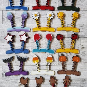 May include: A set of 16 wooden spring-loaded headbands with various holiday and seasonal themes. The headbands are painted in bright colors and feature designs such as snowmen, hearts, shamrocks, flowers, stars, balloons, pumpkins, candy corn, turkeys, and gingerbread men.