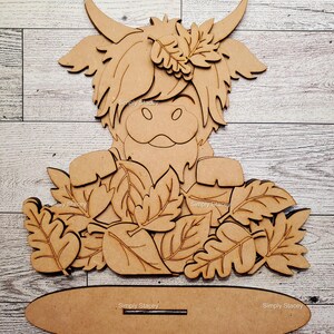 May include: A wooden cutout of a Highland cow with a leaf crown, surrounded by leaves. The cutout is on a wooden base with the text "Simply Stacey" printed on it.