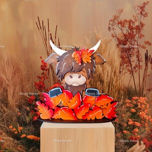 May include: A wooden Highland Cow figurine with a brown and black fur pattern, wearing a crown of orange and red fall leaves. The figurine is standing on a black base and surrounded by orange and red leaves.