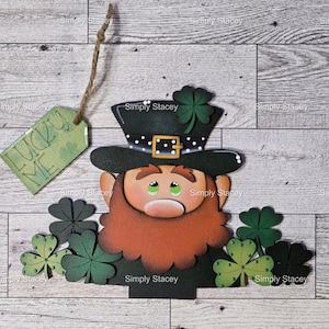 May include: A St. Patrick's Day decoration featuring a leprechaun with a green hat and red beard, surrounded by shamrocks. A tag with the words "LUCKY ME" hangs from the top. The design is on a wooden background.