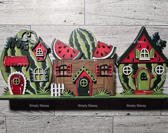 May include: Three watermelon-shaped houses with red roofs and green trim. The houses are decorated with white windows and doors. The houses are sitting on a wooden shelf with a green grass background. The houses are decorated with the words "Simply Stacey".