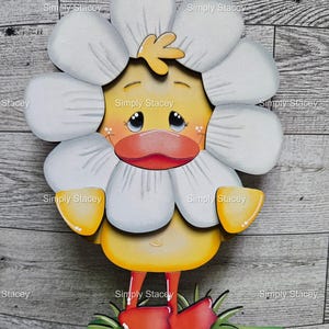 May include: A wooden, whimsical duck figure with a daisy-like flower head. The duck has a yellow body, orange feet, and red shoes. The figure is on a green base with grass and the text "Simply Stacey".