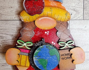 May include: A wooden gnome decoration with a red body, a rainbow hat, and a globe. The gnome features an apple with the words "A TEACHER" and a ruler with the number "3". It also has the words "HOME WORK".