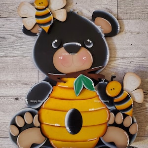 May include: A black bear figurine holding a yellow and black beehive with a green leaf. The bear has a brown nose and white eyes. There are two bees on either side of the bear.