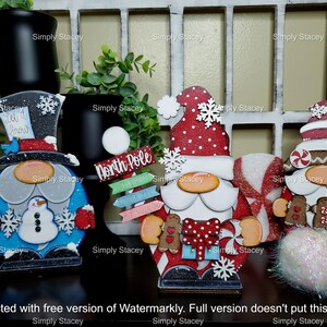 May include: Four wooden Christmas gnomes with different designs. The gnome on the left is wearing a blue hat with a snowman on his belly. The gnome in the middle is wearing a red hat with white polka dots and holding a candy cane. The gnome on the right is wearing a brown hat with a candy cane and gingerbread cookies. The gnomes are all standing on a wooden surface.