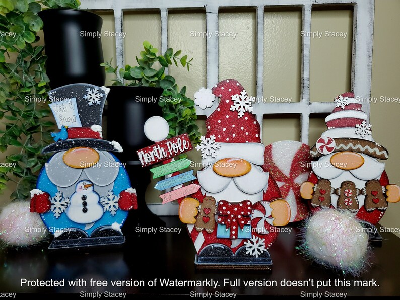 May include: Four wooden Christmas gnomes with different designs. The gnome on the left is wearing a blue hat with a snowman on his belly. The gnome in the middle is wearing a red hat with white polka dots and holding a candy cane. The gnome on the right is wearing a brown hat with a candy cane and gingerbread cookies. The gnomes are all standing on a wooden surface.
