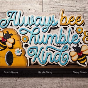 May include: Wooden sign with the text "Always bee humble & kind" in blue and yellow lettering. The sign features a beehive with a bee on each side.