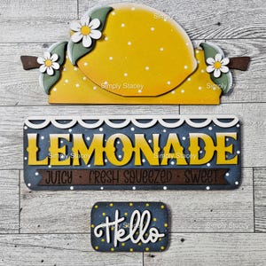 May include: Wooden signs with a lemon design and the word "LEMONADE". The lemon sign is yellow with white polka dots and daisy accents. The "LEMONADE" sign is blue with yellow lettering and the words "JUICY FRESH SQUEEZED SWEET". A small sign says "Hello".