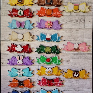 May include: A set of 15 wooden bows with various painted designs. The bows are decorated with different themes, including a sunflower, a pumpkin, a Christmas tree, a butterfly, a cupcake, a maple leaf, a shamrock, a bunny, a watermelon, a snowflake, a daisy, an apple, a spider, a heart, a star, and a sun.