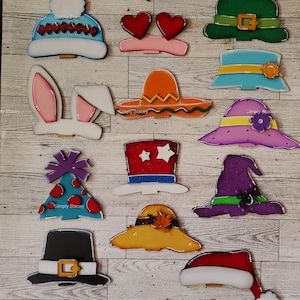 May include: A set of 12 wooden door hangers, each featuring a different hat design. The hats include a Santa hat, a witch hat, a top hat, a bunny ear headband, a party hat, a sombrero, a leprechaun hat, a heart headband, a pilgrim hat, a fall hat, a winter hat, and a blue hat with a flower.