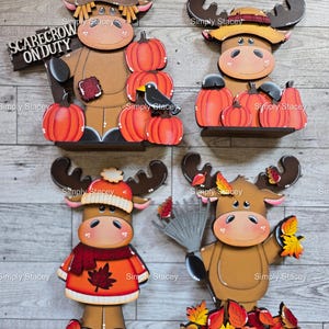 May include: Four wooden moose figurines with fall themes. One is a scarecrow holding a sign that reads "Scarecrow on Duty". Other moose are decorated with pumpkins, leaves, and hats. The moose are brown, orange, and red.