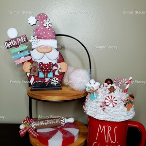 May include: A tiered tray decorated for Christmas with a wooden Santa figurine, a red mug with white lettering that says "Mrs. Claus", and a gingerbread-themed decoration. The tray is decorated with white and red candy canes and a white fluffy pom-pom.
