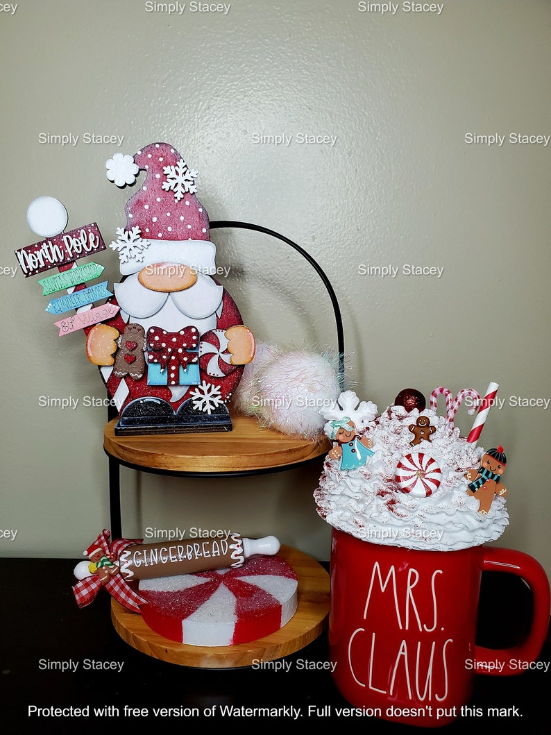 May include: A tiered tray decorated for Christmas with a wooden Santa figurine, a red mug with white lettering that says "Mrs. Claus", and a gingerbread-themed decoration. The tray is decorated with white and red candy canes and a white fluffy pom-pom.