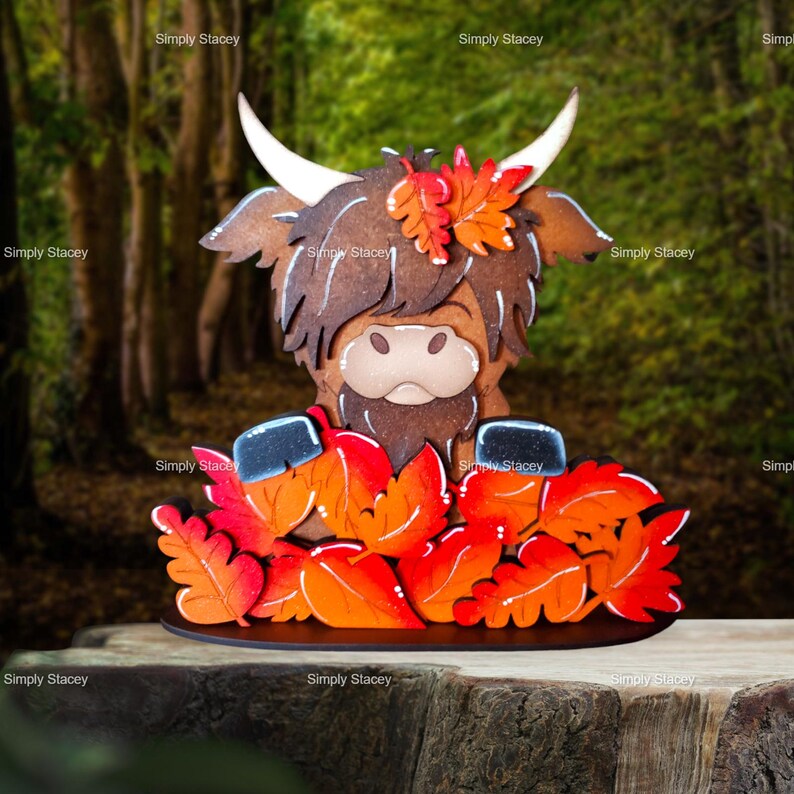 May include: A wooden figurine of a Highland cow with a brown and white face, wearing a crown of orange and red fall leaves. The cow is standing on a black base with a brown wooden surface behind it.