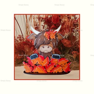 May include: A wooden Highland cow figurine with a brown and white fur pattern, surrounded by orange and red fall leaves. The cow is standing on a wooden base.