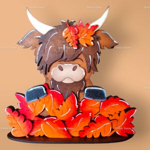 May include: A wooden Highland Cow figurine with brown fur and a black nose, surrounded by orange and red painted leaves. The figurine is standing on a black base.
