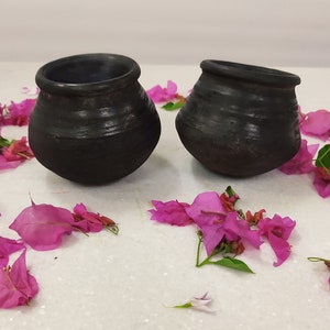 Puede incluir: Dos ollas de barro negro con una superficie rugosa y texturizada. Las ollas est&aacute;n sobre una superficie blanca con p&eacute;talos de flores rosas esparcidos a su alrededor.