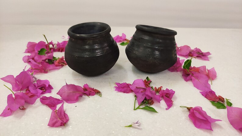 Puede incluir: Dos ollas de barro negro con una superficie rugosa y texturizada. Las ollas est&aacute;n sobre una superficie blanca con p&eacute;talos de flores rosas esparcidos a su alrededor.