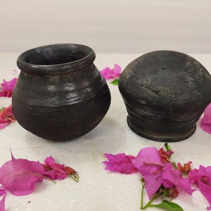 Puede incluir: Dos ollas de cer&aacute;mica negra, una con tapa redonda y la otra con una abertura ancha, se muestran sobre una superficie blanca rodeada de p&eacute;talos de flores rosas.