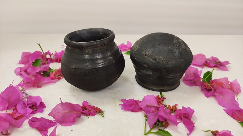 Puede incluir: Dos ollas de cer&aacute;mica negra, una con tapa redonda y la otra con una abertura ancha, se muestran sobre una superficie blanca rodeada de p&eacute;talos de flores rosas.
