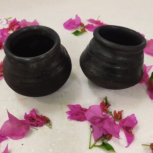 Puede incluir: Dos ollas de barro negro con una superficie rugosa y texturizada. Las ollas est&aacute;n sobre una superficie blanca con p&eacute;talos de flores rosas esparcidos a su alrededor.