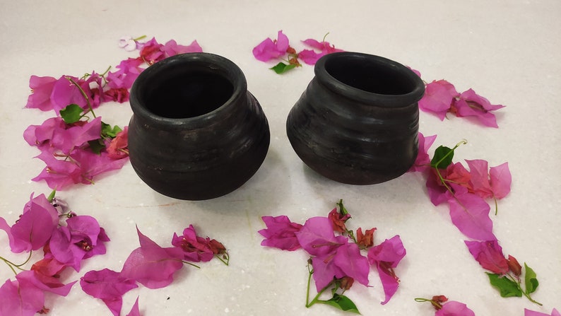 Puede incluir: Dos ollas de barro negro con una superficie rugosa y texturizada. Las ollas est&aacute;n sobre una superficie blanca con p&eacute;talos de flores rosas esparcidos a su alrededor.