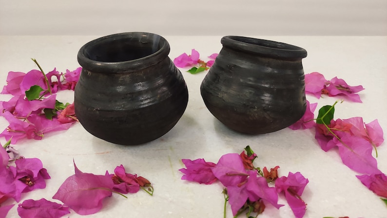 Puede incluir: Dos ollas de barro negro con una superficie rugosa y texturizada. Las ollas est&aacute;n sobre una superficie blanca con p&eacute;talos de flores rosas esparcidos a su alrededor.