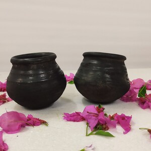 Puede incluir: Dos ollas de barro negro con una superficie rugosa y texturizada, sentadas sobre una superficie blanca con p&eacute;talos de flores rosas esparcidos a su alrededor.