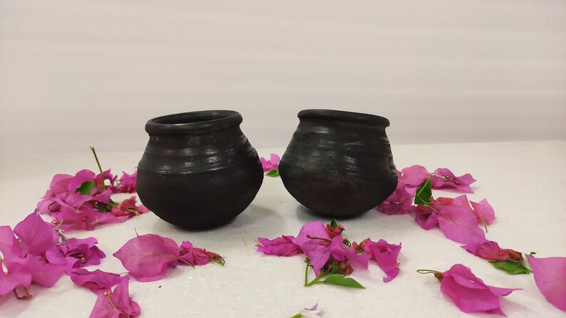 Puede incluir: Dos ollas de barro negro con una superficie rugosa y texturizada, sentadas sobre una superficie blanca con p&eacute;talos de flores rosas esparcidos a su alrededor.