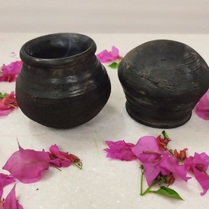 Puede incluir: Dos macetas de cer&aacute;mica negra, una con tapa redonda y la otra una maceta m&aacute;s alta y redonda, se muestran sobre una superficie blanca rodeada de p&eacute;talos de flores rosas.