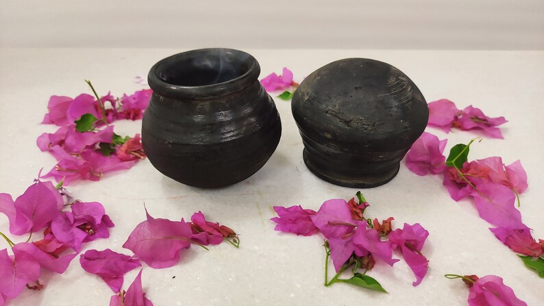 Puede incluir: Dos macetas de cer&aacute;mica negra, una con tapa redonda y la otra una maceta m&aacute;s alta y redonda, se muestran sobre una superficie blanca rodeada de p&eacute;talos de flores rosas.