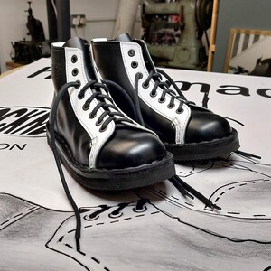 May include: A pair of black and white leather boots with white laces. The boots have a thick black sole and are tied at the ankle.