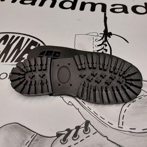 Puede incluir: Primer plano de una suela de goma negra con un patrón de banda de rodadura texturizado. La suela está diseñada para una bota y tiene un pequeño agujero ovalado en el centro.