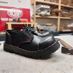 以下が含まれることがあります： 赤いステッチと厚いソールが付いた黒い革靴。靴は灰色の表面にあり、背景にはハンマーとドライバーがあります。