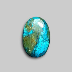 May include: An oval-shaped gemstone with a polished surface. The stone features a blend of turquoise, teal, and olive green hues, with a marbled appearance. The gemstone is set against a neutral gray background.